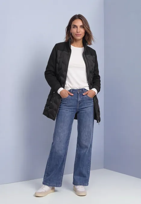 Frau in langer gesteppter schwarzer Jacke, weißem T-Shirt, weiten Jeans und weißen Sneakern vor schlichtem Hintergrund.