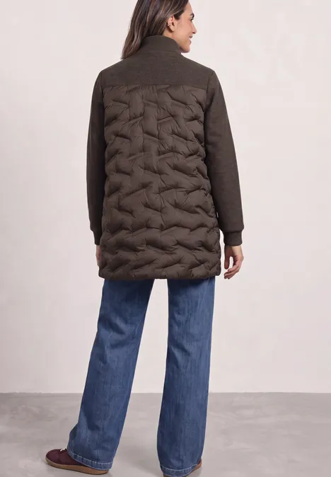 Eine Frau von hinten in dunklem Steppmantel mit Zopfmuster auf dem Rücken, weite Jeans und Schuhe.