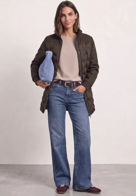 Frau in olivgrünem Steppmantel, beigem Pullover, ausgestellten Jeans, bordeauxfarbigen Loafers und hellblauer Tasche.