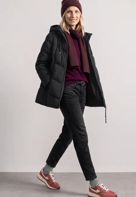 Femme en doudoune noire, bonnet et écharpe bordeaux, jean et baskets, souriante en marchant.