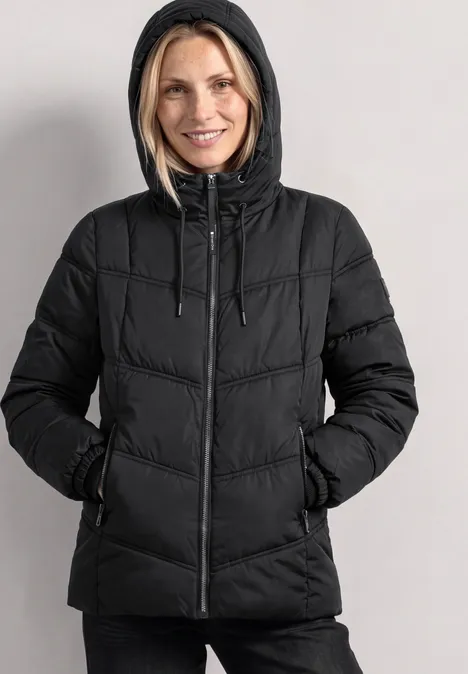 Femme portant une doudoune noire matelassée à capuche, mains dans les poches, sourire aux lèvres.