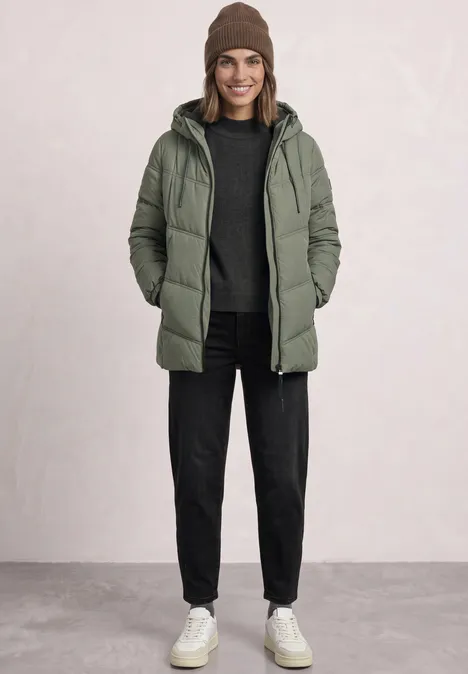 Eine Frau in grüner Steppjacke, schwarzer Hose, Mütze und weißen Sneakers steht vor hellem Hintergrund.