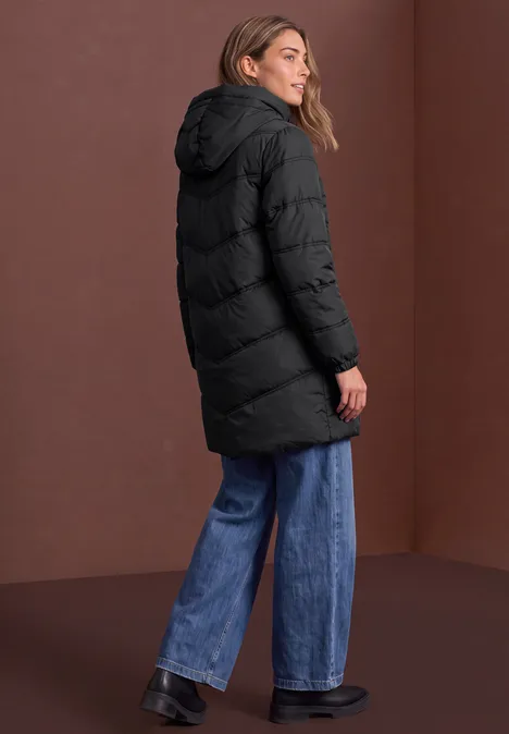 Persoon in lange zwarte gewatteerde jas met capuchon, wijde jeans en donkere schoenen voor een effen achtergrond.