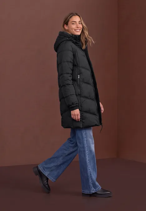 Vrouw met zwarte gewatteerde jas, blauwe spijkerbroek en zwarte schoenen poseert zijwaarts voor een bruine achtergrond.