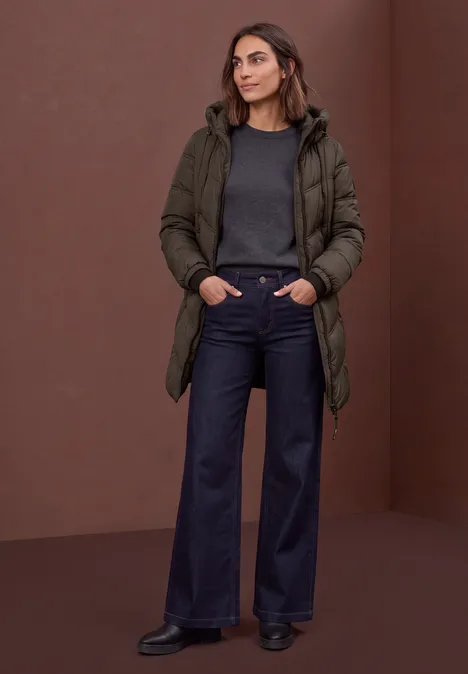 Een vrouw in een donkergroene winterjas, grijze trui en wijde jeans voor een bruine achtergrond.