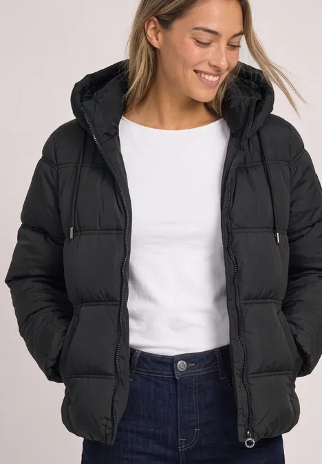 Frau in schwarzer Steppjacke mit Kapuze, weißem T-Shirt und Jeans, lächelnd mit Händen in den Taschen.