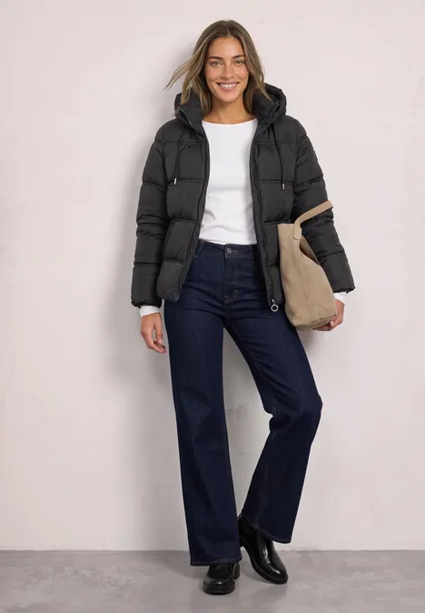 Eine lächelnde Frau trägt eine schwarze Steppjacke, ein weißes Shirt, weite Jeans und eine Tasche.