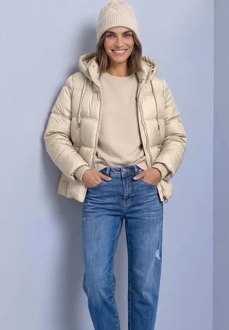 Veste matelassée à capuche barley beige Une femme sourit devant un fond clair, portant une doudoune beige, un bonnet, un pull et un jean bleu.