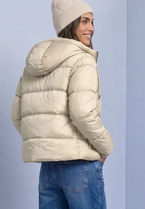 Veste matelassée à capuche barley beige Femme porte une doudoune beige claire avec capuche et bonnet, regard par-dessus l'épaule en souriant.