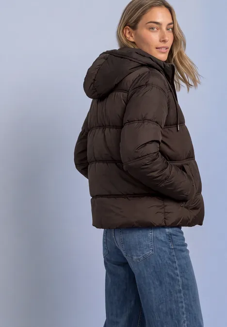 Une femme porte une doudoune marron à capuche et un jean bleu devant un fond clair.