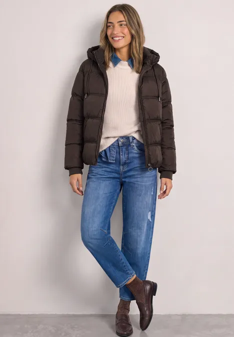 Femme souriante en doudoune sombre, pull clair, jean et bottines marron devant un fond neutre.