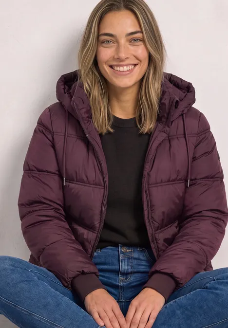 Une femme souriante assise jambes croisées, portant une doudoune violette, un pull noir et un jean.