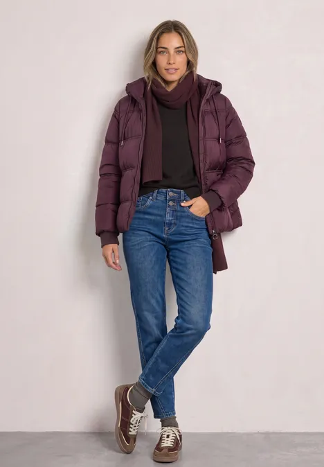 Femme en doudoune violette, pull sombre, jean bleu et baskets, posant décontractée devant un mur.