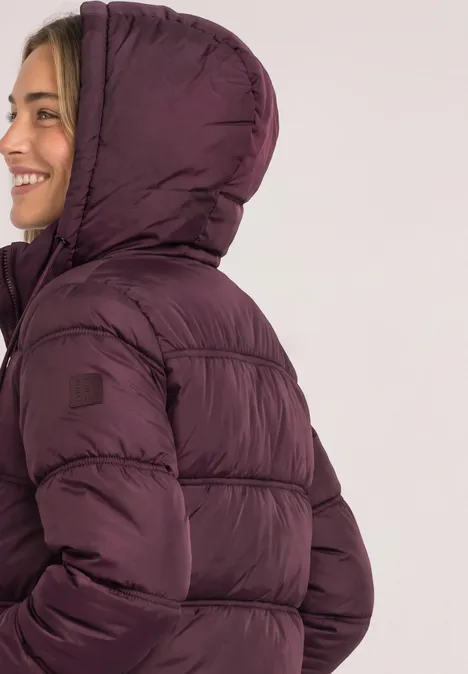 Femme de profil portant une doudoune violette à capuche, souriant vers la gauche.