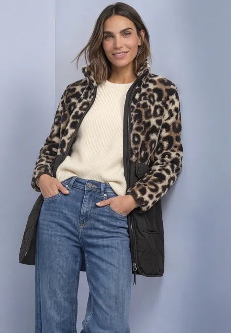 Eine Frau mit Leopardenjacke, weißem Pullover und Jeans steht lächelnd vor hellem Hintergrund.