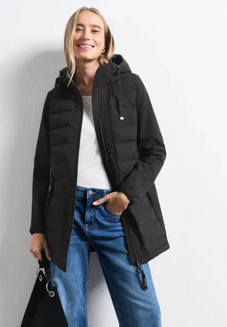 Frau trägt schwarzen Parka mit Kapuze, weiße Bluse und blaue Jeans, schaut lächelnd in die Kamera.