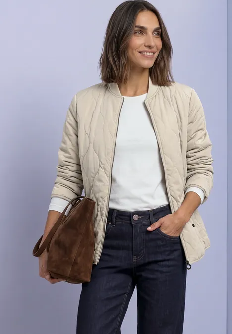 Frau in beiger Steppjacke, weißem T-Shirt und blauen Jeans, hält eine braune Wildledertasche.