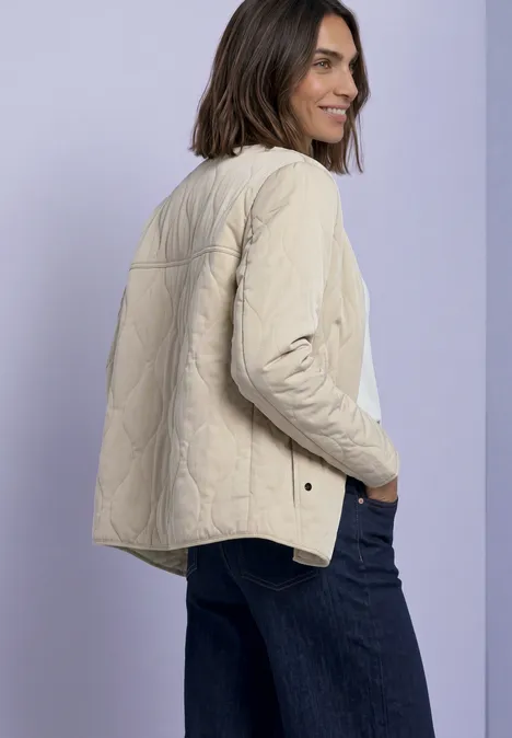 Frau mit schulterlangem Haar trägt eine beige Steppjacke und blaue Jeans, steht vor hellem Hintergrund und lächelt.