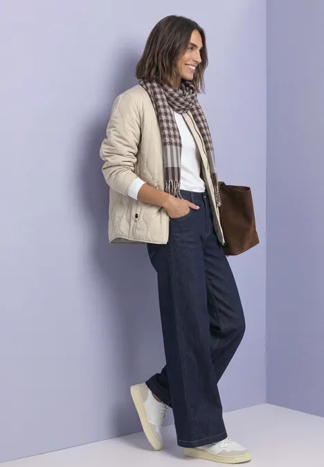 Eine Frau steht vor lila Wand, trägt beige Jacke, karierten Schal, weite Jeans, weiße Sneaker und braune Tasche.