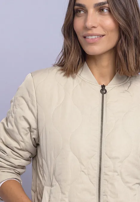 Frau trägt eine beige, gesteppte Jacke mit Reißverschluss und Rundhalsausschnitt vor hellem Hintergrund.