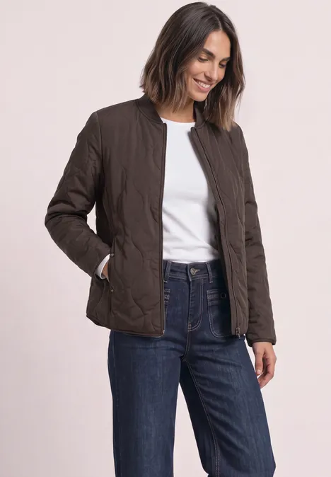 Frau trägt eine Steppjacke in Dunkelbraun, dazu weißes Shirt und Jeans, sie lächelt.