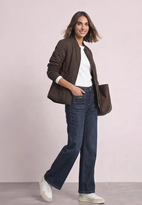 Frau in brauner Steppjacke, weißem T-Shirt, weiten Jeans und weißen Sneakern, mit Tasche vor hellem Hintergrund.