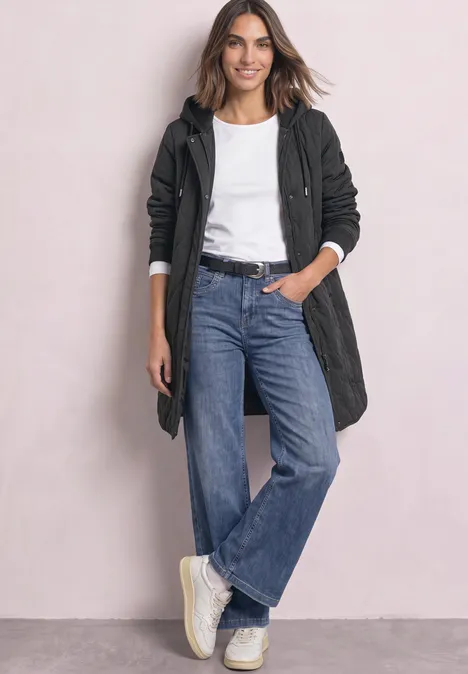 Frau in schwarzem Mantel, weißem T-Shirt, weitem Jeans und Sneakern vor hellem Hintergrund.