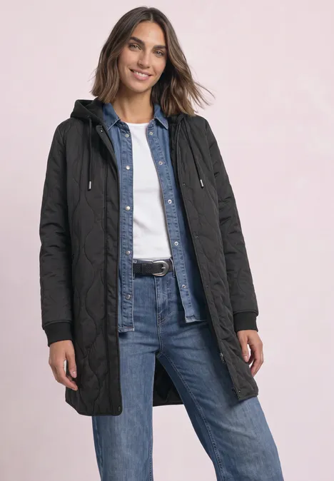 Frau trägt gesteppte schwarze Jacke mit Kapuze, darüber Jeanshemd und weißes Shirt, kombiniert mit blauen Jeans.