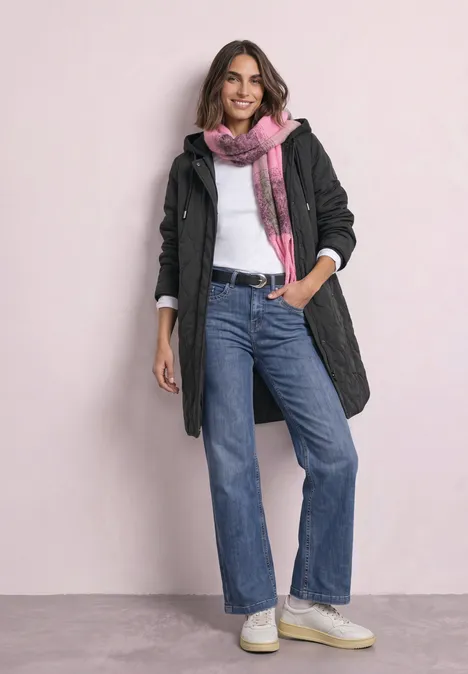Eine Frau in dunklem Mantel, Jeans, rosafarbenem Schal und weißen Schuhen steht vor hellem Hintergrund und lächelt.