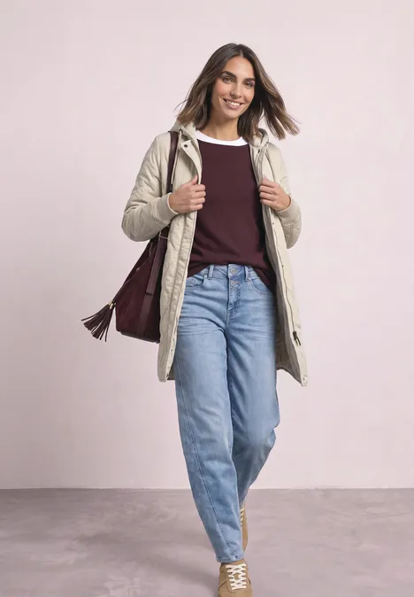 Une femme souriante en jean, pull et manteau, marche avec un sac en bandoulière.