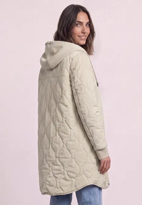 Femme en manteau matelassé long beige à capuche, vue de côté, souriante.