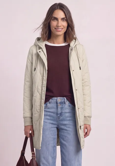 Une femme porte un parka clair, un haut bordeaux et un jean, tenant un sac à main marron.