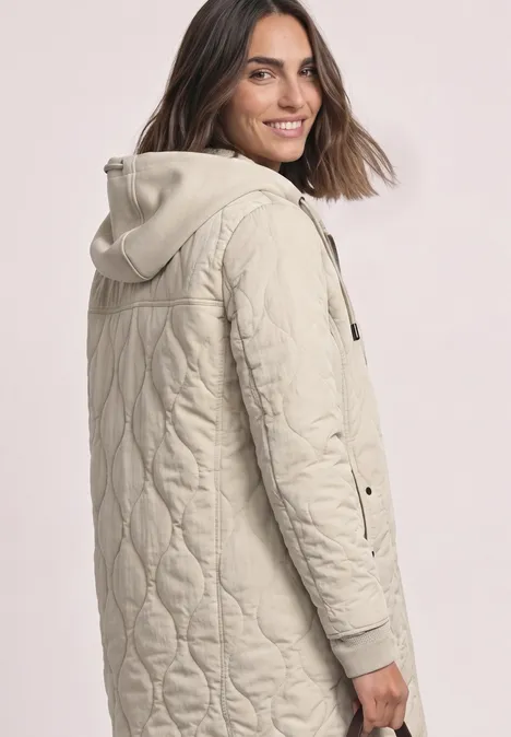 Une femme souriante porte une veste matelassée à capuche beige clair, avec des boutons-pression sur les côtés.