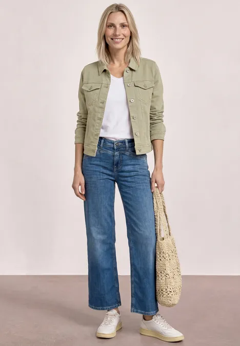 Frau trägt hellgrüne Jacke, weißes Shirt, blaue Jeans und helle Sneaker, mit geflochtener Tasche.