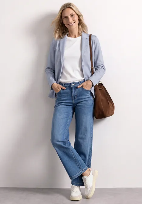 Basic Piqué Blazer whispery blue Leicht verschwommene Frau an der Wand, hellblauer Blazer, weißes Shirt, gerade Jeans, weiße Sneaker, braune Tasche.