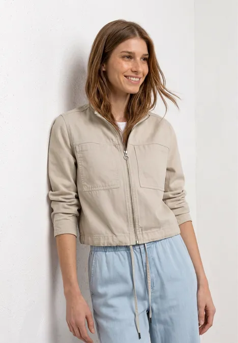 Een vrouw voor een witte achtergrond draagt een zandkleurig jasje met rits en een losse lichtblauwe broek.