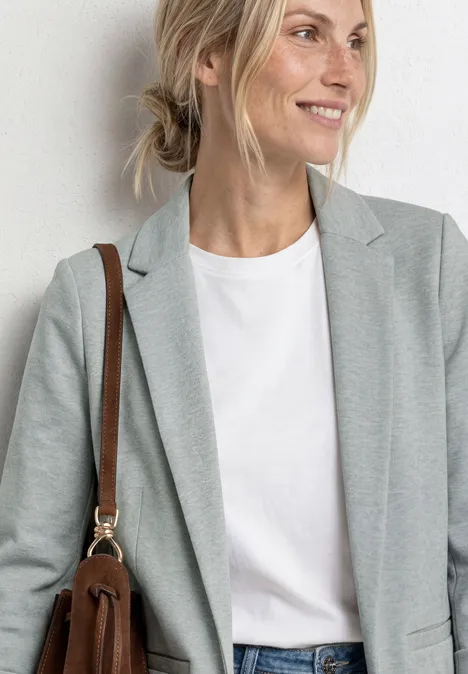 Frau in grauem Blazer, weißem T-Shirt und brauner Schultertasche vor neutralem Hintergrund.