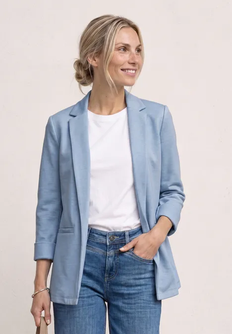 Frau trägt hellblauen Blazer, weißes Shirt und Jeans, steht lässig mit einer Hand in der Tasche.