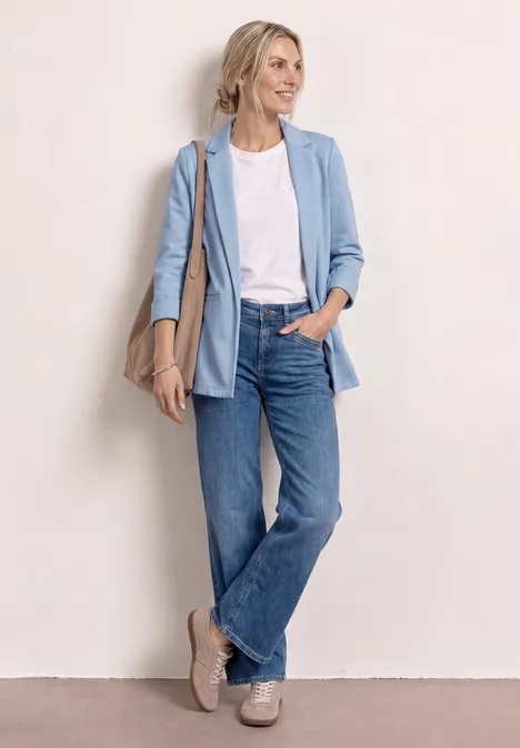 Frau in hellem Blazer, weißem Shirt, blauen Jeans, Sneakers und Schultertasche.