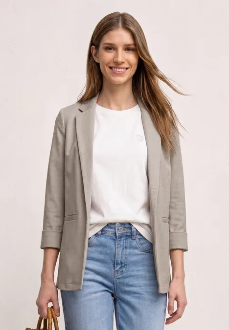 Frau in weißem T-Shirt, grauem Blazer und blauen Jeans vor hellem Hintergrund