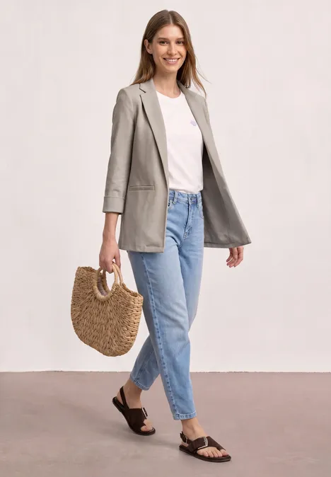 Frau trägt grauen Blazer, weißes Shirt, blaue Jeans, schwarze Sandalen und hält eine Korbtasche.
