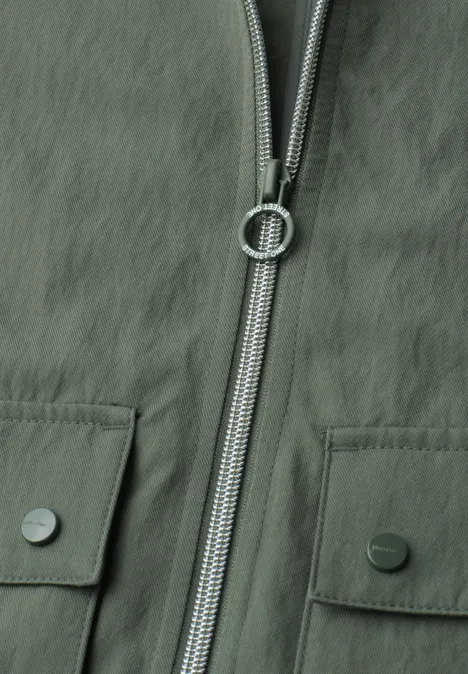 Nahaufnahme einer olivgrünen Jacke mit Reißverschluss und zwei aufgesetzten Taschen mit Metallknöpfen.