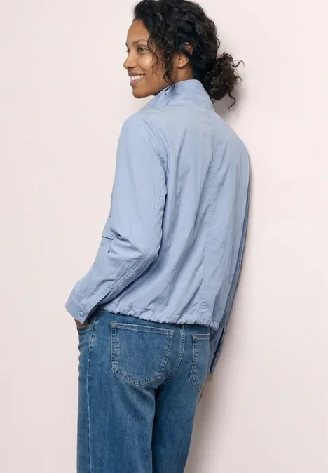 Person von hinten in hellblauer Bluse und Jeans vor hellem Hintergrund, dunkel gelockte Haare im Dutt.