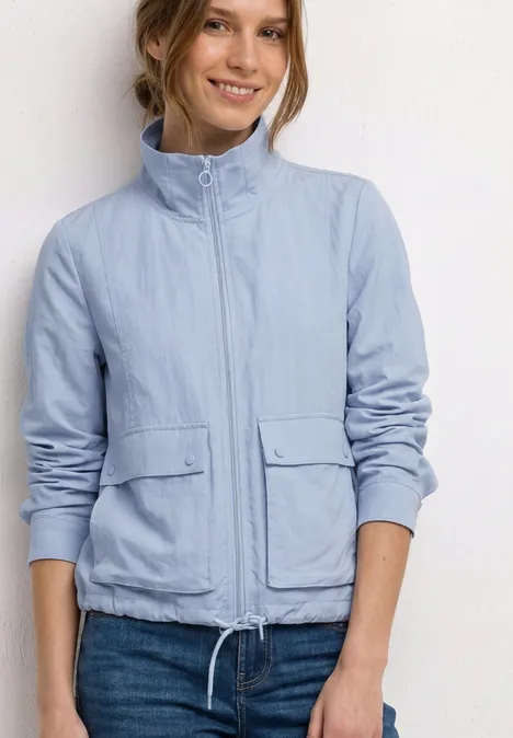 Person mit hellblauer Jacke und blauer Jeans vor schlichtem weißen Hintergrund.