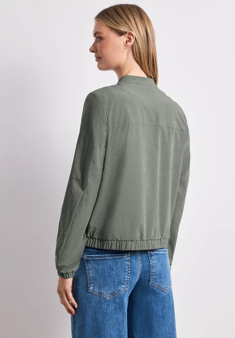 Frau von hinten mit olivgrünem Blouson und blauen Jeans vor hellem Hintergrund.