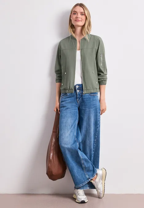 Person in grüner Jacke, weißem Top und weiten Jeans, mit brauner Tasche und Sneakern vor weißer Wand.