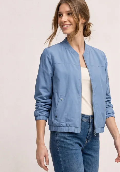 Eine Frau in heller Jeansjacke, weißem Top und blauer Jeans vor hellem Hintergrund, Gesicht unkenntlich.