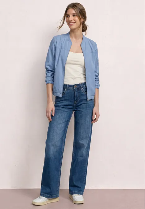 Eine Frau trägt eine hellblaue Bomberjacke, weißes Top, hochgeschnittene weite Jeans und weiße Sneaker.