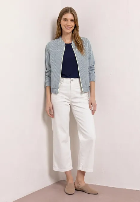 Vrouw in lichtblauw jasje, donkerblauw topje, witte wijde jeans en beige loafers voor een neutrale achtergrond.