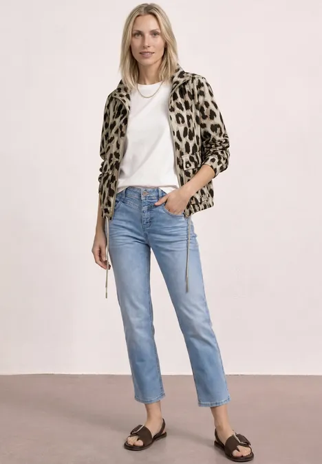 Vrouw in luipaardprintjas, wit t-shirt, rechte blauwe jeans en zwarte sandalen voor een neutrale achtergrond